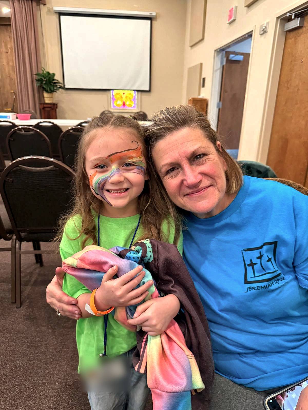 A smiling girl with a rainbow painted on her face hugging a volunteer