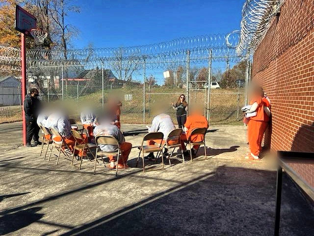 Inmates seated in a circle in the yard while a volunteer speaks; faces are blurred for privacy